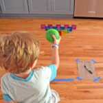 Young child making a bowling game out of a green rubber ball and magnetic tiles.