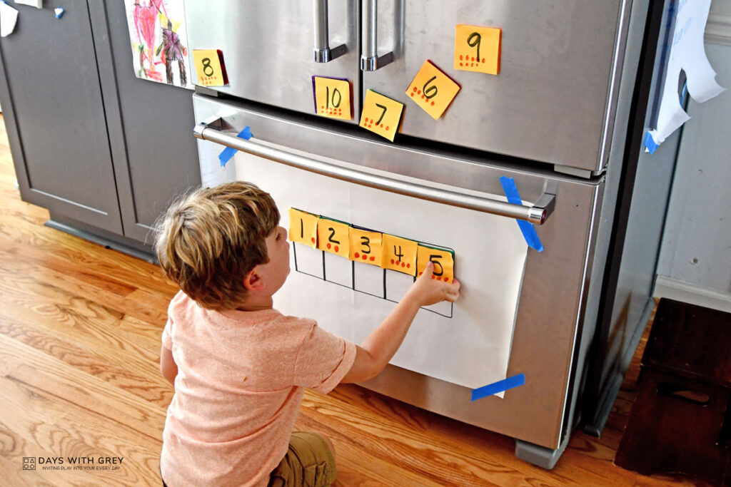 Preschooler adding the number five to his ten frame that is drawn on the lower level of the refrigerator.
