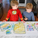 Two preschoolers at the table are painting winter hat drawings with watercolor paints and smiling.