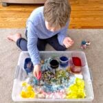 Boy adding red colored water into a sensory bin filled with snow.