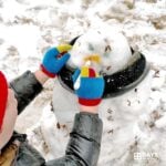 Young child using an orange marker outside in the snow to make a carrot nose for his snowman.