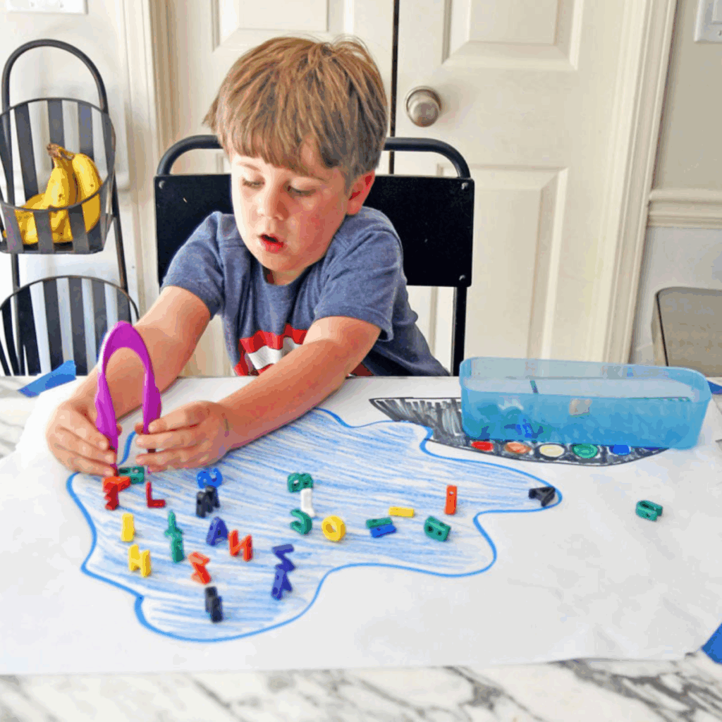 Preschool boy using tongs to collect lacing letters that are in a drawn pond to help rescue them.