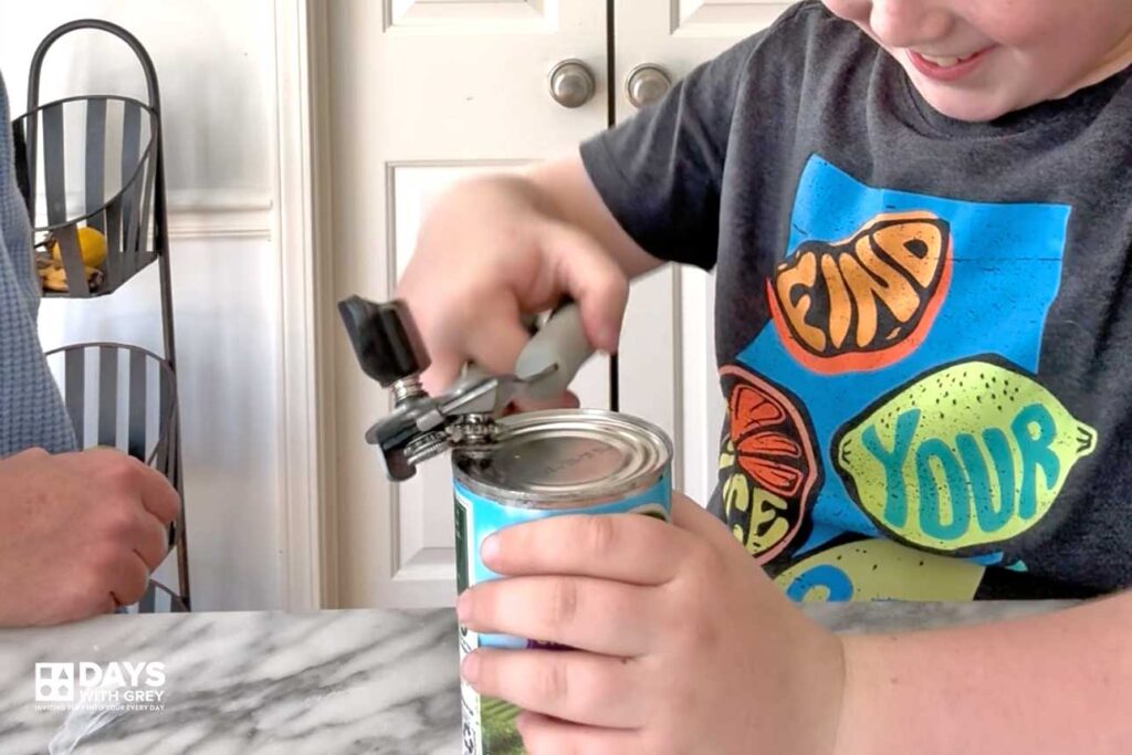 A ten year old boy learning how to use a can opener and opening the can himself.