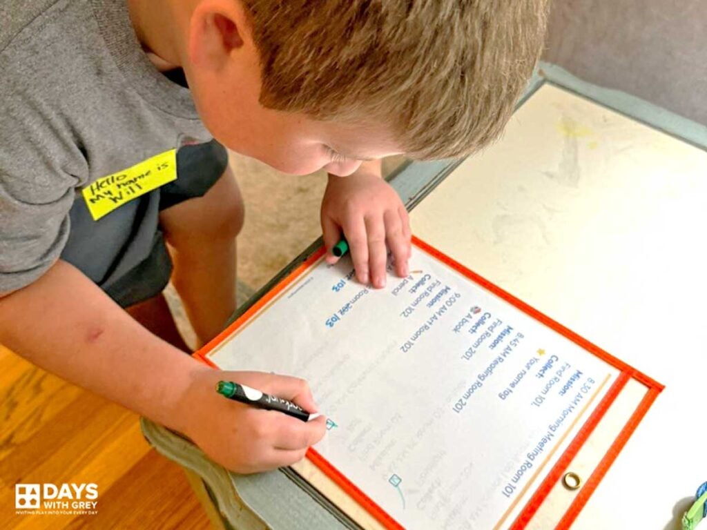 A young boy making a checkmark off of his pretend back to school schedule. The house is marked with room numbers and he is pretending to follow his schedule finding the different rooms.